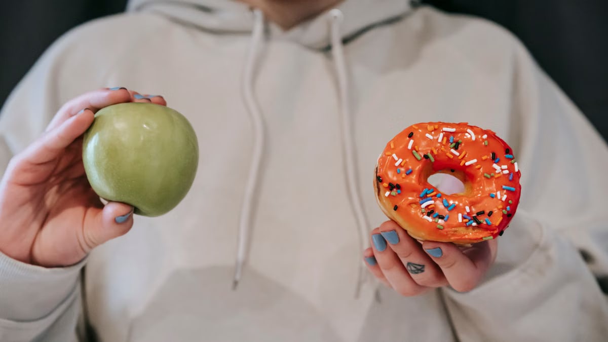 Crop unrecognizable female in loose hoodie showing healthy green apple and sweet delicious donut